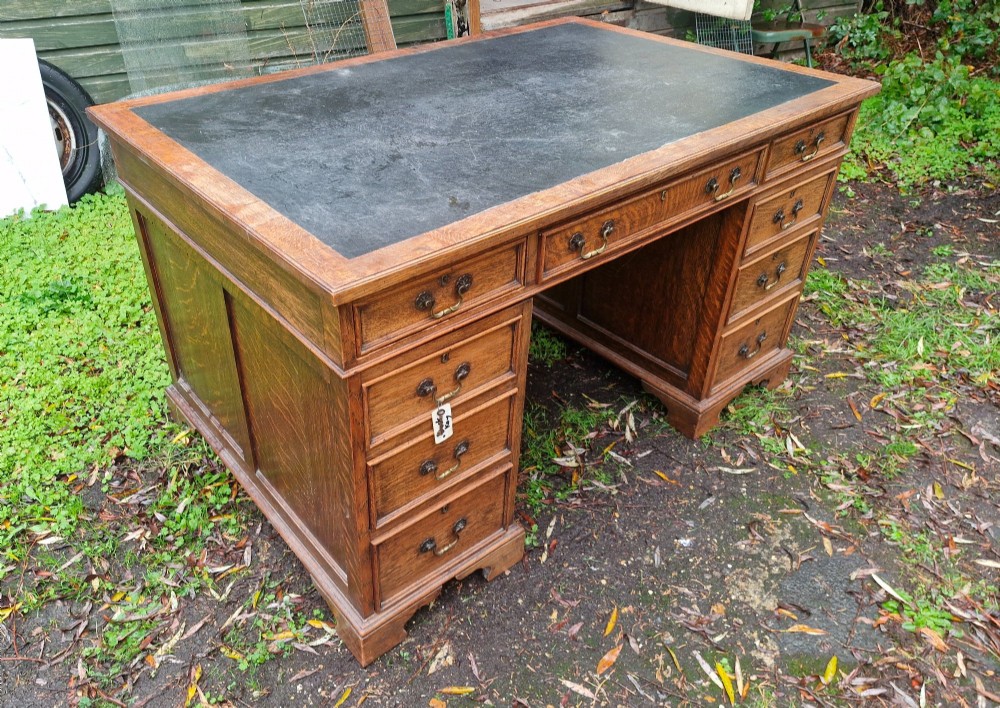 An antique oak pedestal writing desk featuring a black inset leather writing surface, multiple drawers with brass drop handles, and panelled sides. View IV