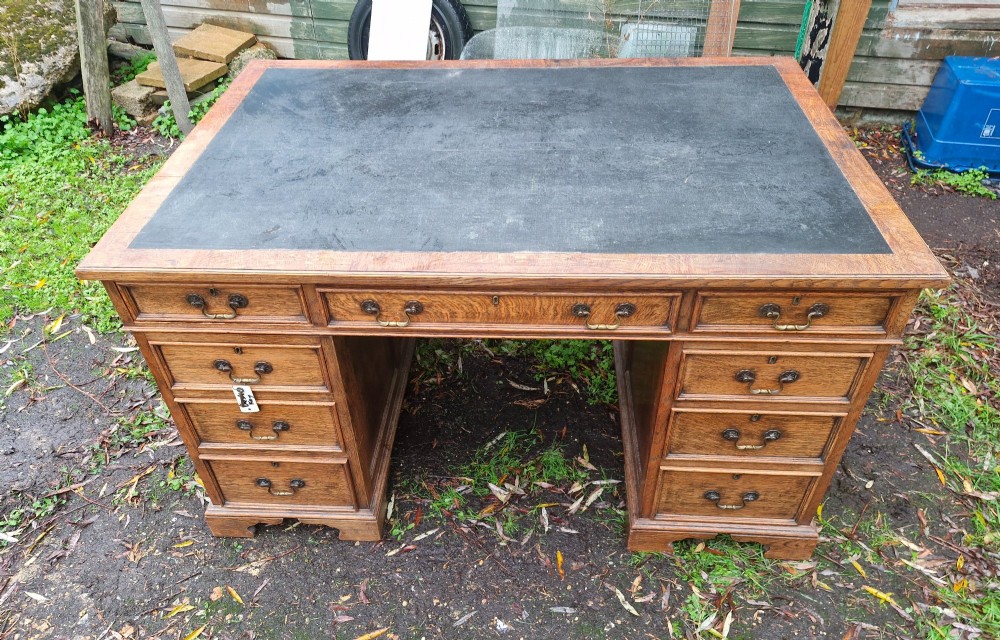 An antique oak pedestal writing desk featuring a black inset leather writing surface, multiple drawers with brass drop handles, and panelled sides. View V