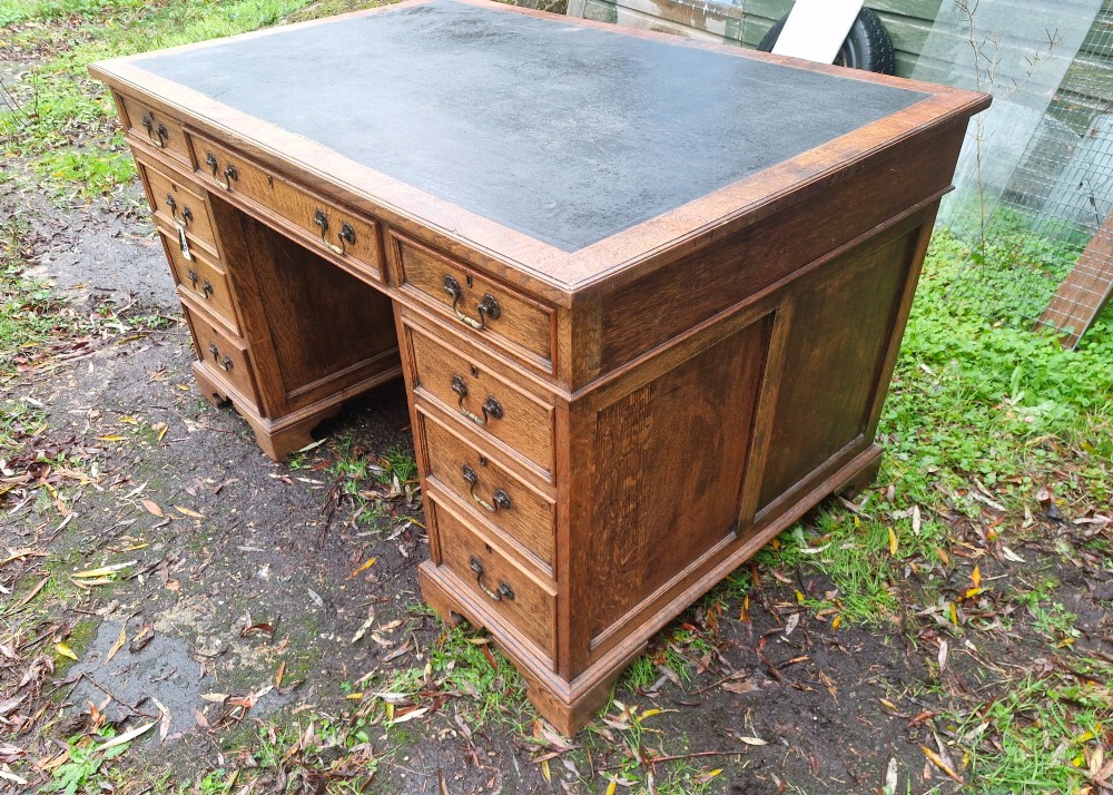 An antique oak pedestal writing desk featuring a black inset leather writing surface, multiple drawers with brass drop handles, and panelled sides. View VI