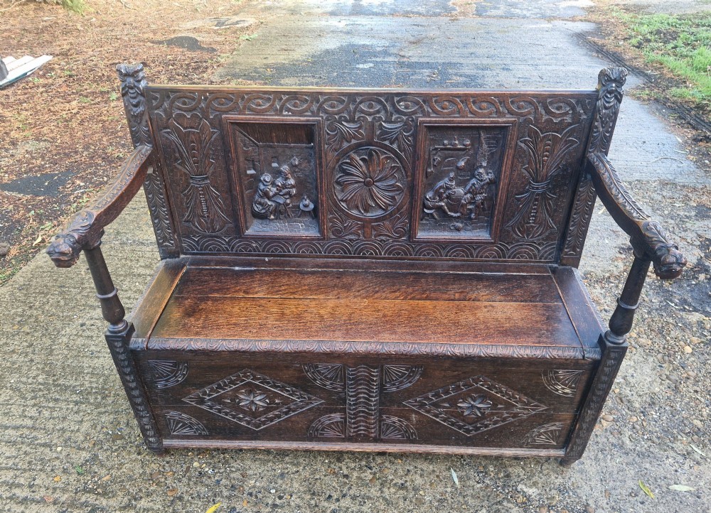 A dark carved oak hall bench featuring a raised back with carved figural and botanical panels, carved armrests, and a hinged storage box base.