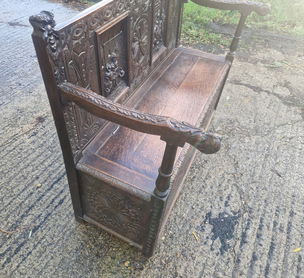 A dark carved oak hall bench featuring a raised back with carved figural and botanical panels, carved armrests, and a hinged storage box base. View III