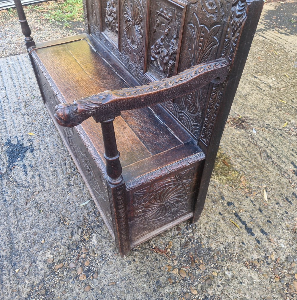 A dark carved oak hall bench featuring a raised back with carved figural and botanical panels, carved armrests, and a hinged storage box base. View IV
