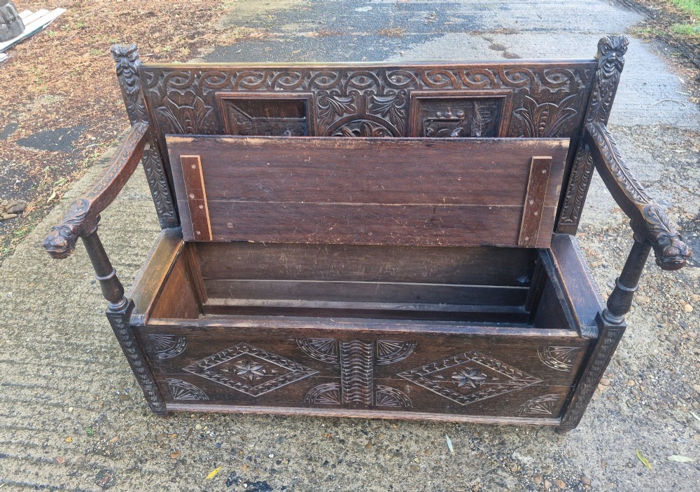 A dark carved oak hall bench featuring a raised back with carved figural and botanical panels, carved armrests, and a hinged storage box base. View V
