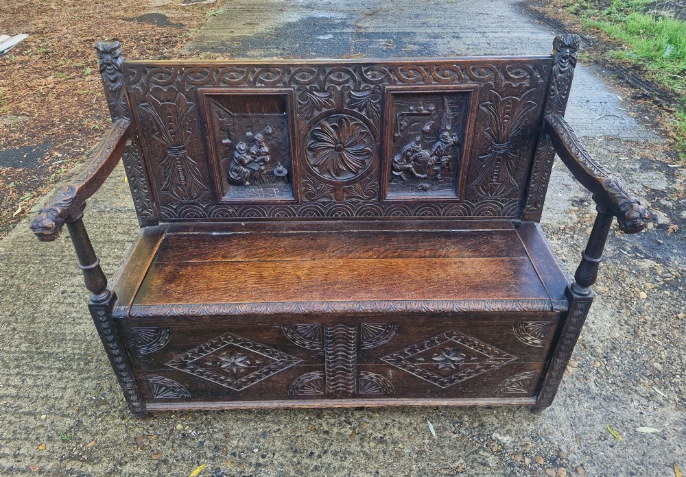 A dark carved oak hall bench featuring a raised back with carved figural and botanical panels, carved armrests, and a hinged storage box base. View VI