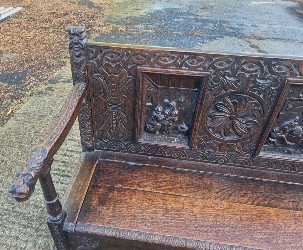 A dark carved oak hall bench featuring a raised back with carved figural and botanical panels, carved armrests, and a hinged storage box base. View VII