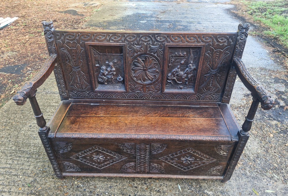 A dark carved oak hall bench featuring a raised back with carved figural and botanical panels, carved armrests, and a hinged storage box base. View VIII
