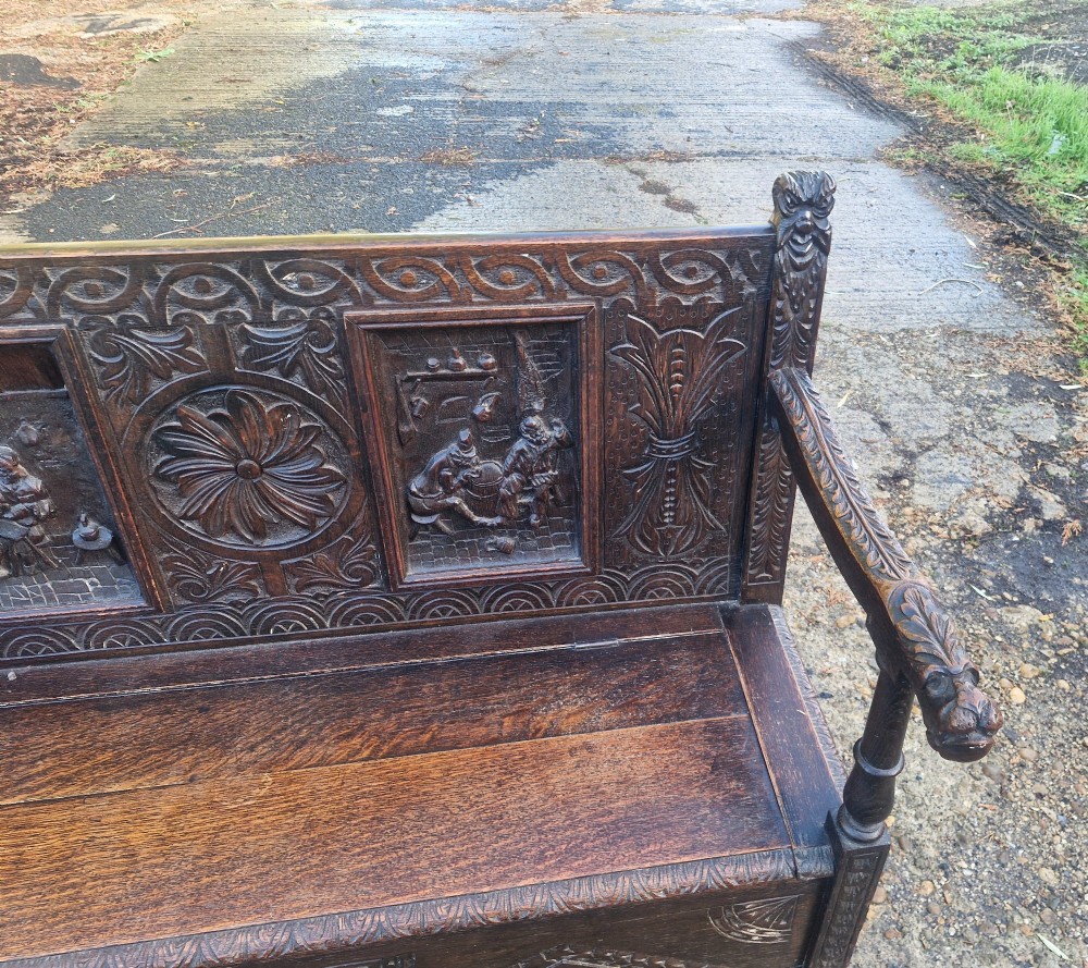 A dark carved oak hall bench featuring a raised back with carved figural and botanical panels, carved armrests, and a hinged storage box base. View IX