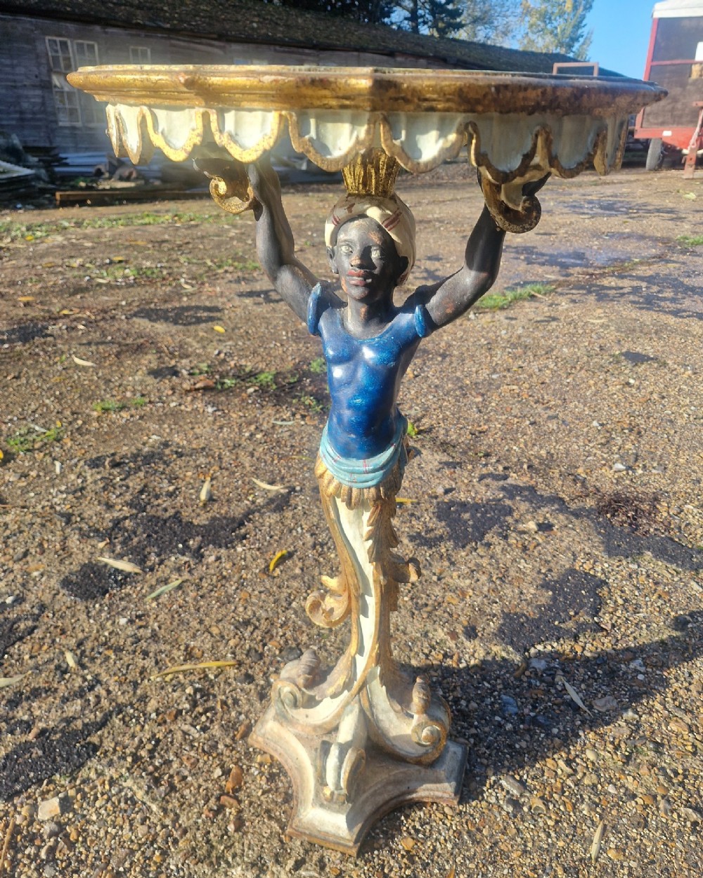A carved and polychrome painted Italian Blackamoor pedestal stand featuring a figure in a blue top supporting a circular scalloped table top over a gilded scroll base. View II