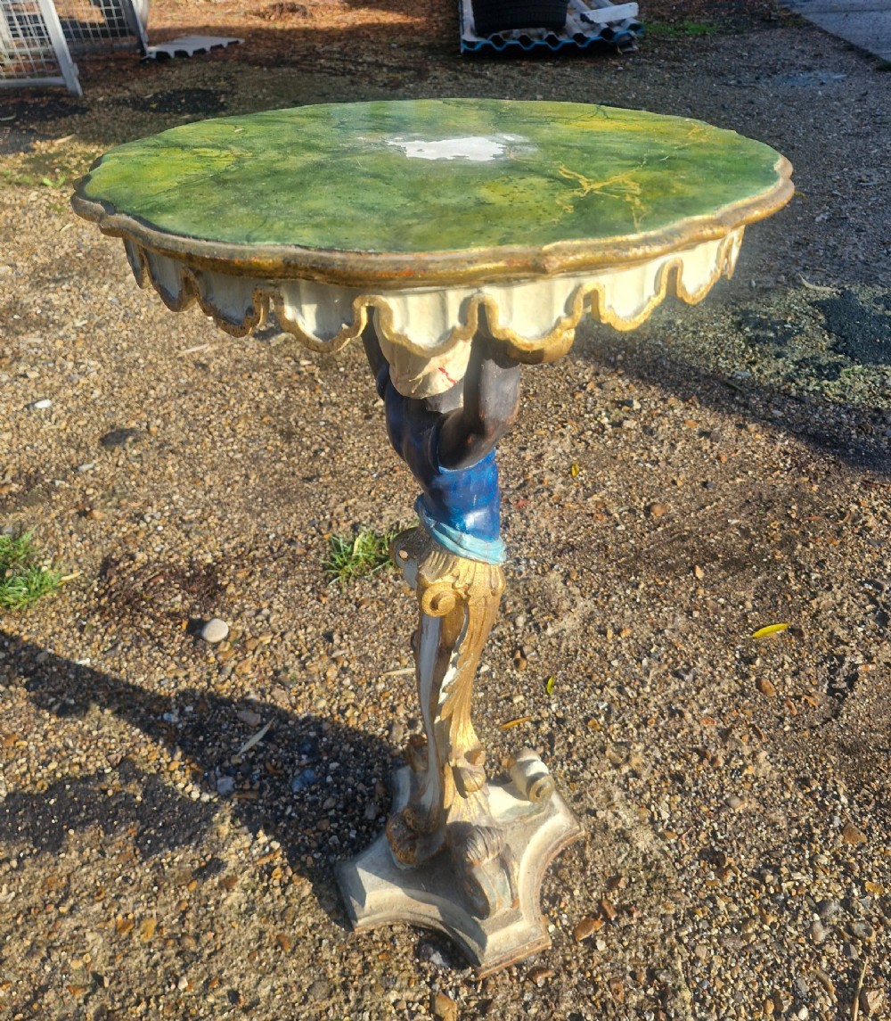 A carved and polychrome painted Italian Blackamoor pedestal stand featuring a figure in a blue top supporting a circular scalloped table top over a gilded scroll base. View V