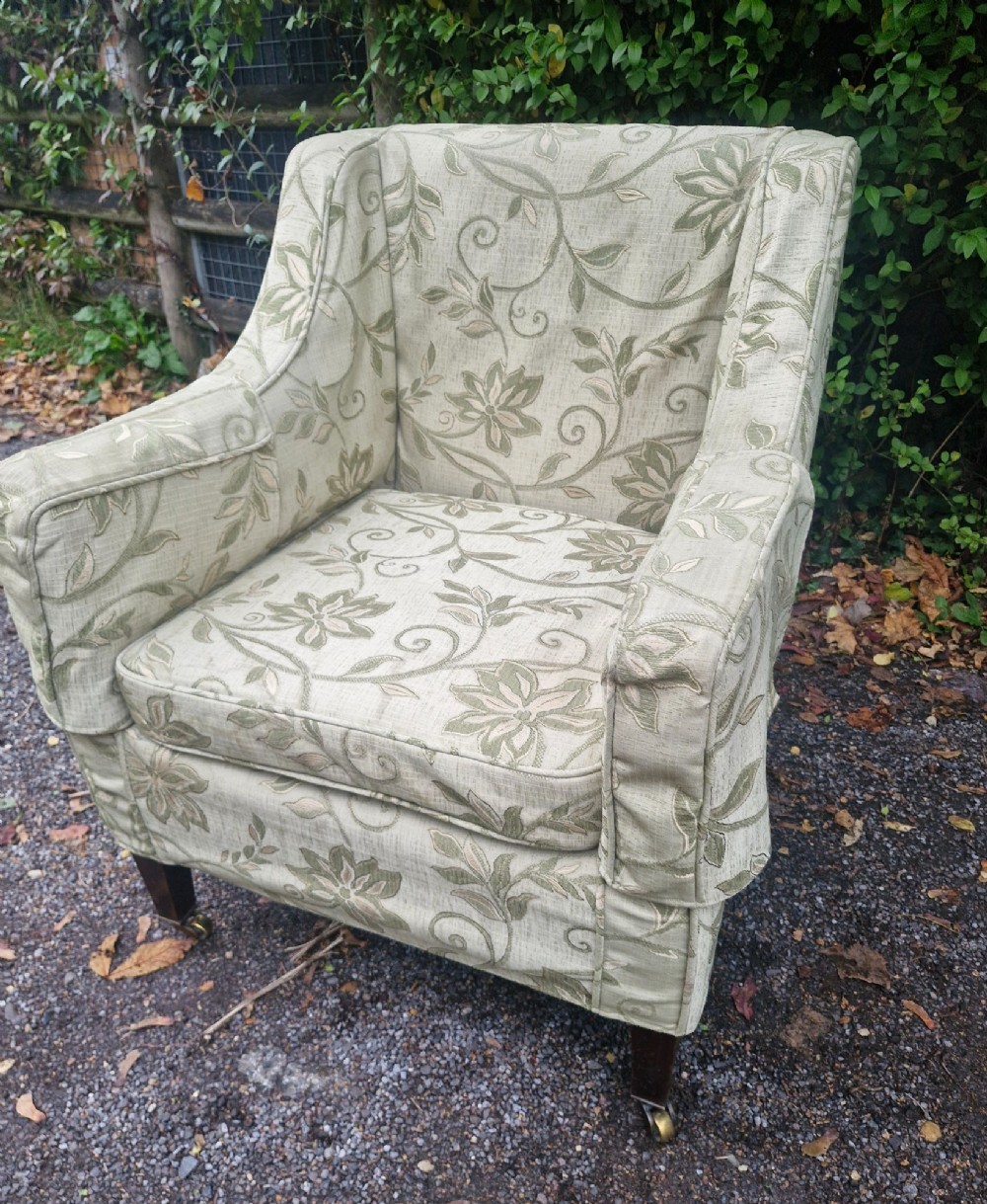 An Edwardian armchair upholstered in patterned green damask fabric with matching braided trim, supported on square tapered mahogany legs with brass castors. View II