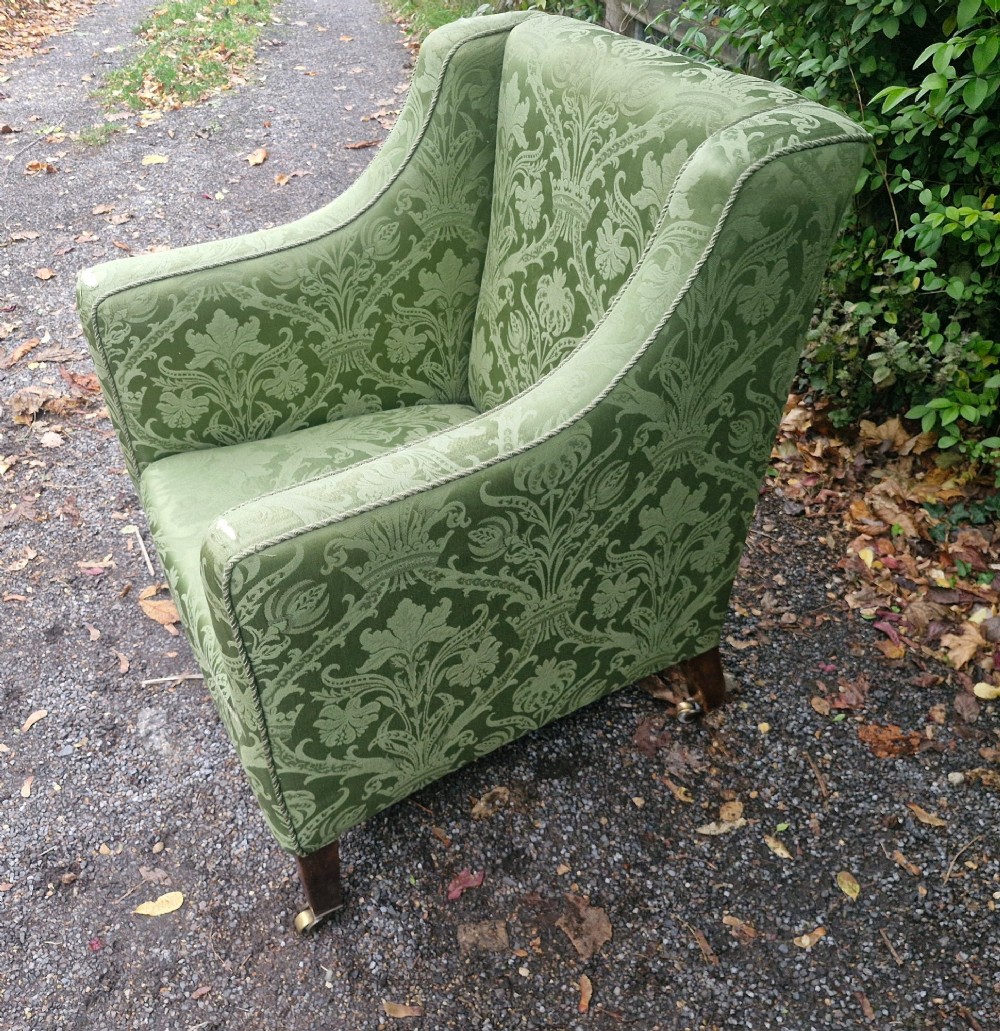 An Edwardian armchair upholstered in patterned green damask fabric with matching braided trim, supported on square tapered mahogany legs with brass castors. View VI