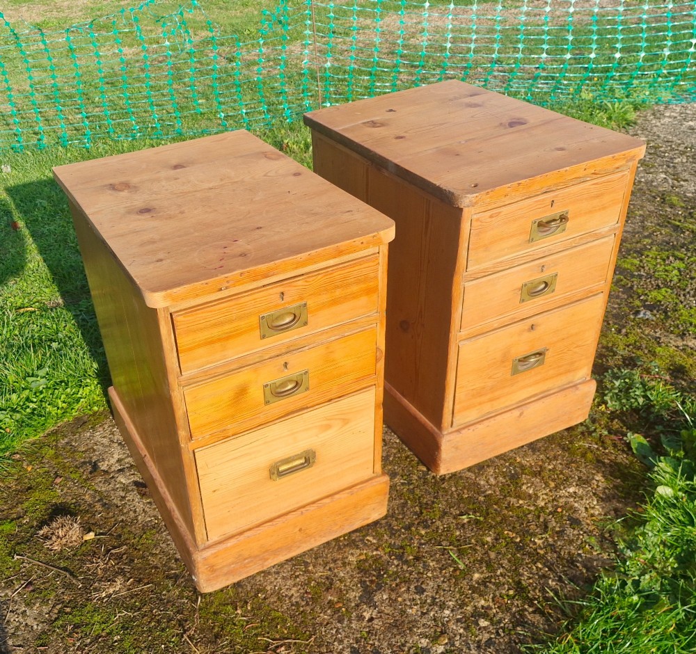 A pair of three-drawer natural pine bedside chests featuring brass flush campaign-style handles and solid plinth bases. View X