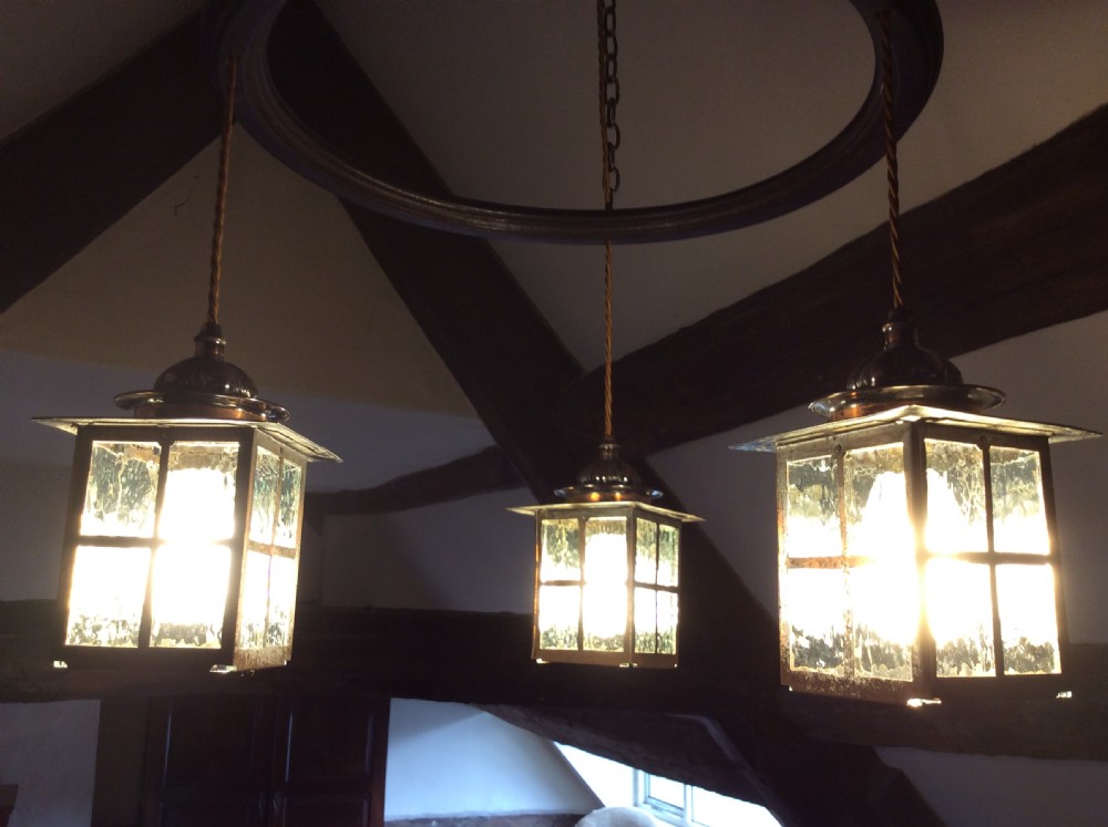 An Arts and Crafts style three-lantern ceiling light featuring a central circular dark frame suspended by chains with three hanging illuminated glass lanterns. View VII