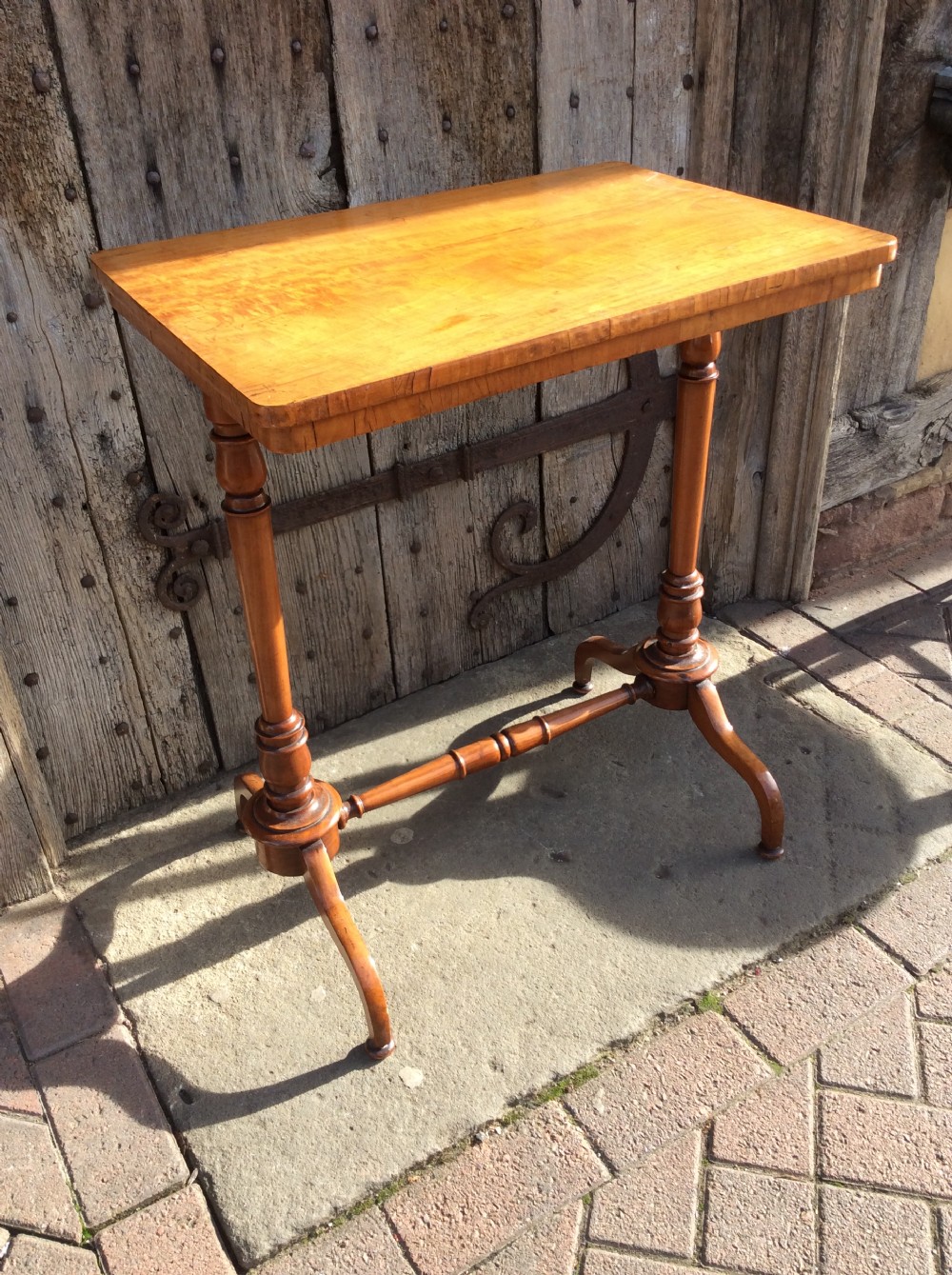 A rectangular 19th-century satinwood lamp table stands outdoors on stone paving against a rustic wooden door, featuring turned uprights and splayed bracket feet. View II