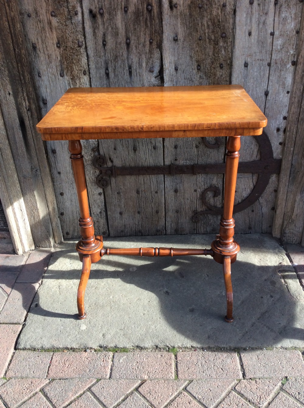 A rectangular 19th-century satinwood lamp table stands outdoors on stone paving against a rustic wooden door, featuring turned uprights and splayed bracket feet. View III