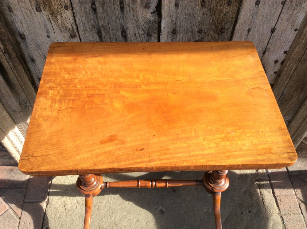 A rectangular 19th-century satinwood lamp table stands outdoors on stone paving against a rustic wooden door, featuring turned uprights and splayed bracket feet. View V