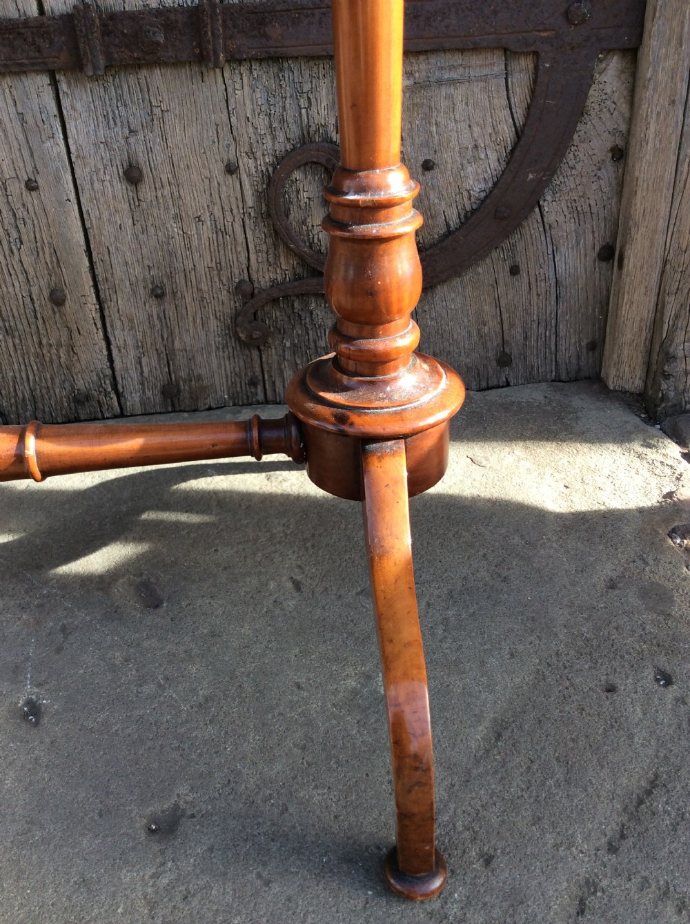 A rectangular 19th-century satinwood lamp table stands outdoors on stone paving against a rustic wooden door, featuring turned uprights and splayed bracket feet. View VI