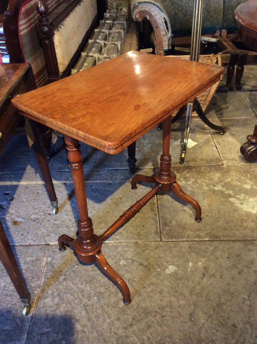 A rectangular 19th-century satinwood lamp table stands outdoors on stone paving against a rustic wooden door, featuring turned uprights and splayed bracket feet. View VII