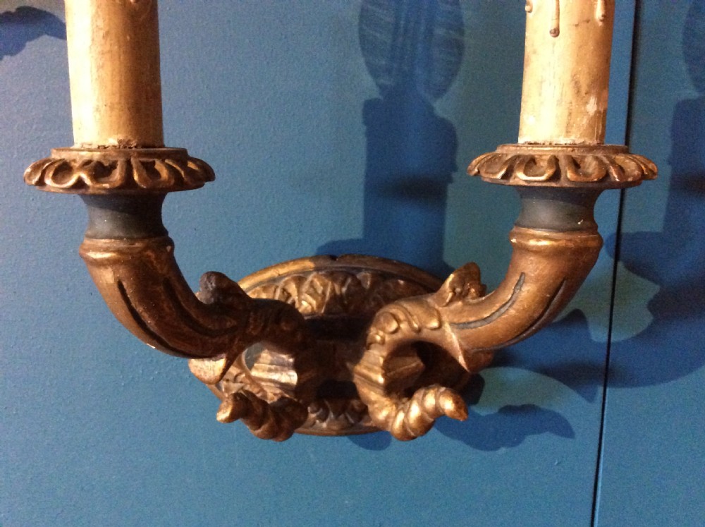 A pair of 19th-century carved gilt-wood twin-arm wall sconces featuring decorative candle sleeves and curved scroll arms mounted on oval backplates. View V
