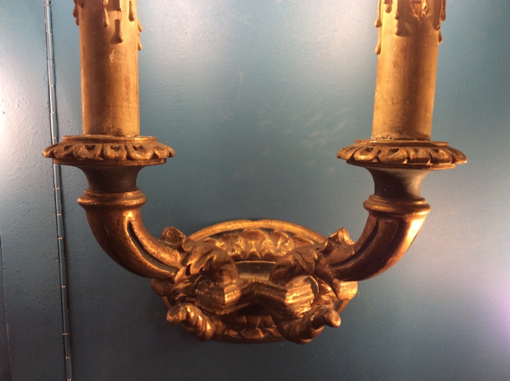 A pair of 19th-century carved gilt-wood twin-arm wall sconces featuring decorative candle sleeves and curved scroll arms mounted on oval backplates. View VI