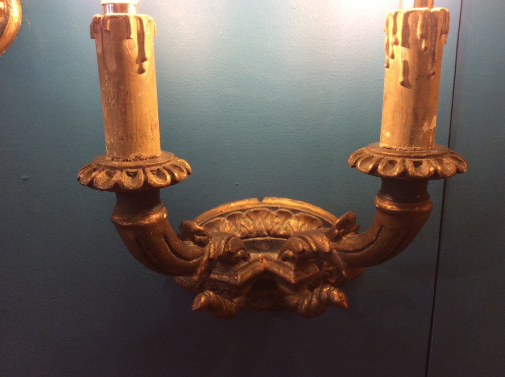 A pair of 19th-century carved gilt-wood twin-arm wall sconces featuring decorative candle sleeves and curved scroll arms mounted on oval backplates. View VII