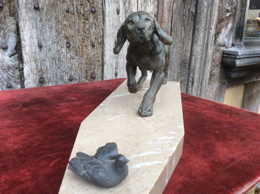 A dark bronze sculpture depicting a hunting dog stalked towards a small bird, mounted on a cream marble plinth. View VI