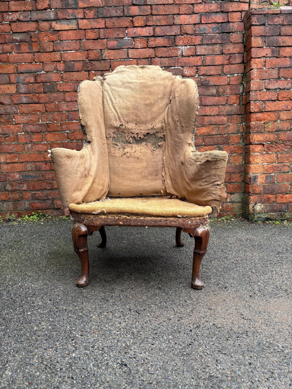 An 18th century Queen Anne wing armchair frame in worn hessian fabric with carved walnut cabriole front legs.