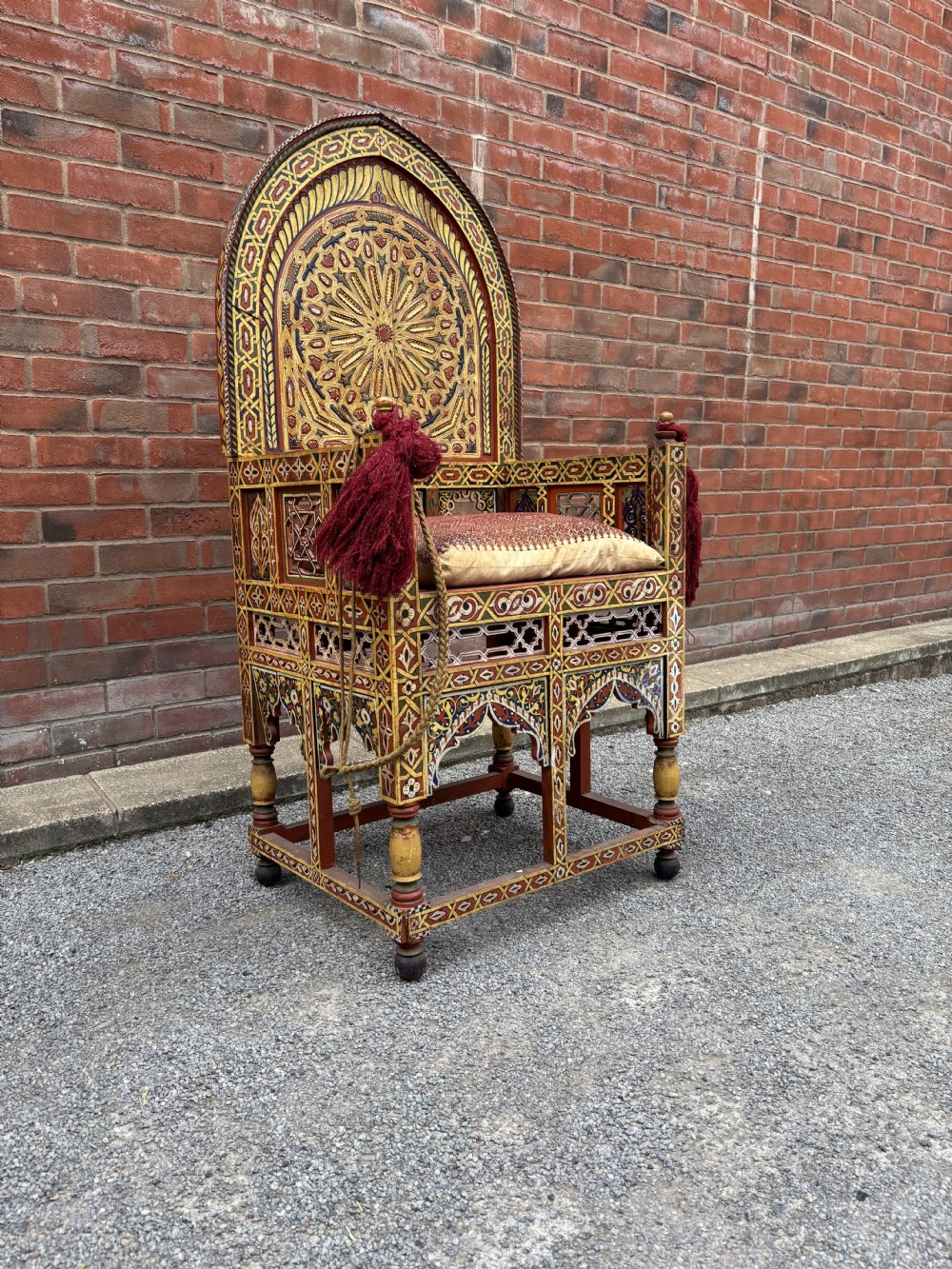 An antique Moroccan wooden armchair featuring an arched backrest intricately hand-painted with geometric starburst motifs in red, yellow, and blue pigments, completed with side tassels and a patterned seat cushion. View II