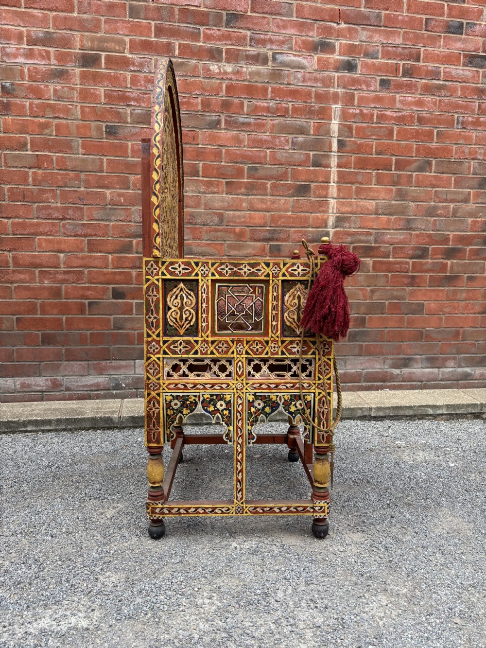 An antique Moroccan wooden armchair featuring an arched backrest intricately hand-painted with geometric starburst motifs in red, yellow, and blue pigments, completed with side tassels and a patterned seat cushion. View III