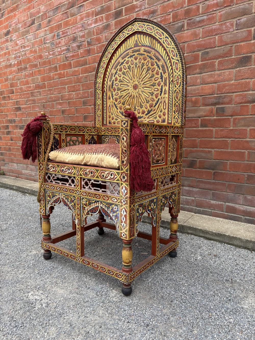An antique Moroccan wooden armchair featuring an arched backrest intricately hand-painted with geometric starburst motifs in red, yellow, and blue pigments, completed with side tassels and a patterned seat cushion. View V
