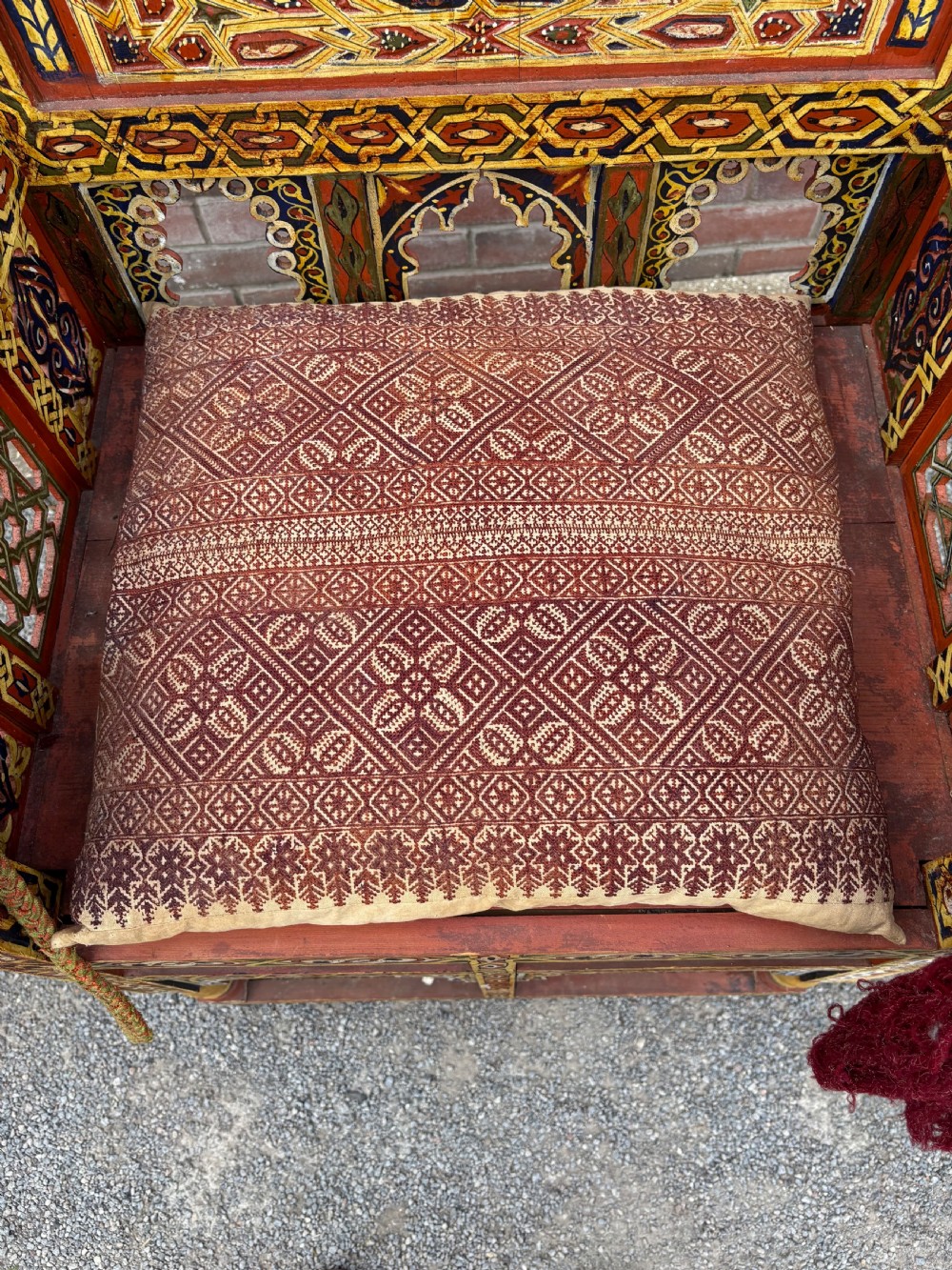 An antique Moroccan wooden armchair featuring an arched backrest intricately hand-painted with geometric starburst motifs in red, yellow, and blue pigments, completed with side tassels and a patterned seat cushion. View VI