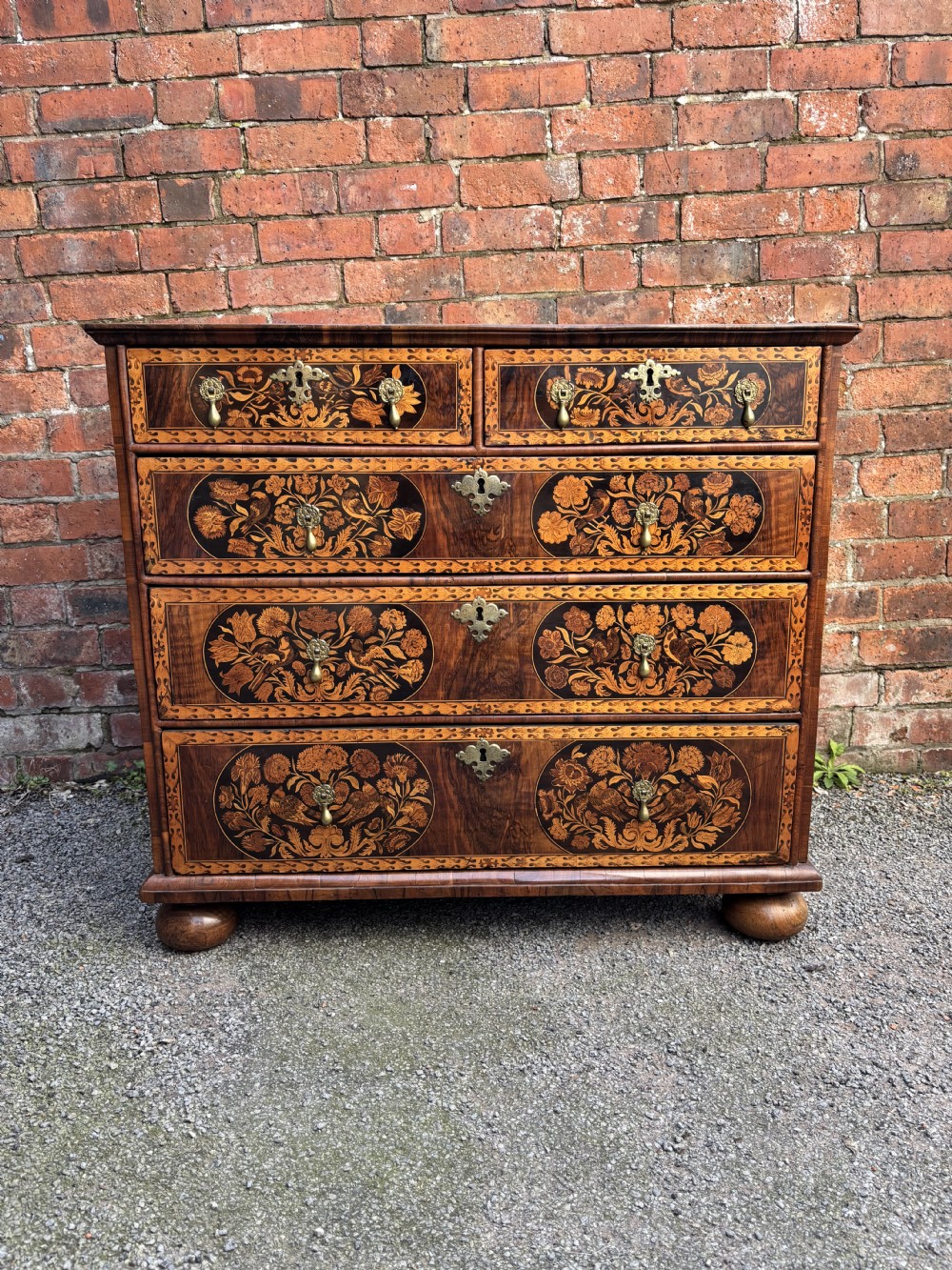 A 17th-century walnut chest of drawers featuring floral marquetry panels, teardrop brass drop handles, and turned bun feet.