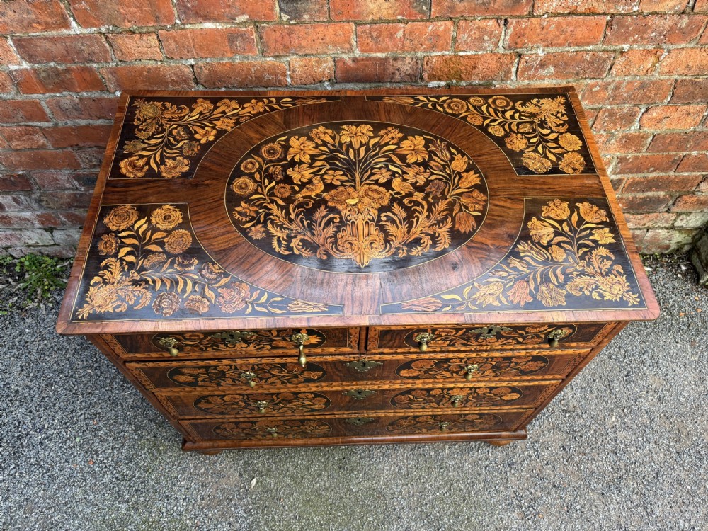 A 17th-century walnut chest of drawers featuring floral marquetry panels, teardrop brass drop handles, and turned bun feet. View VI