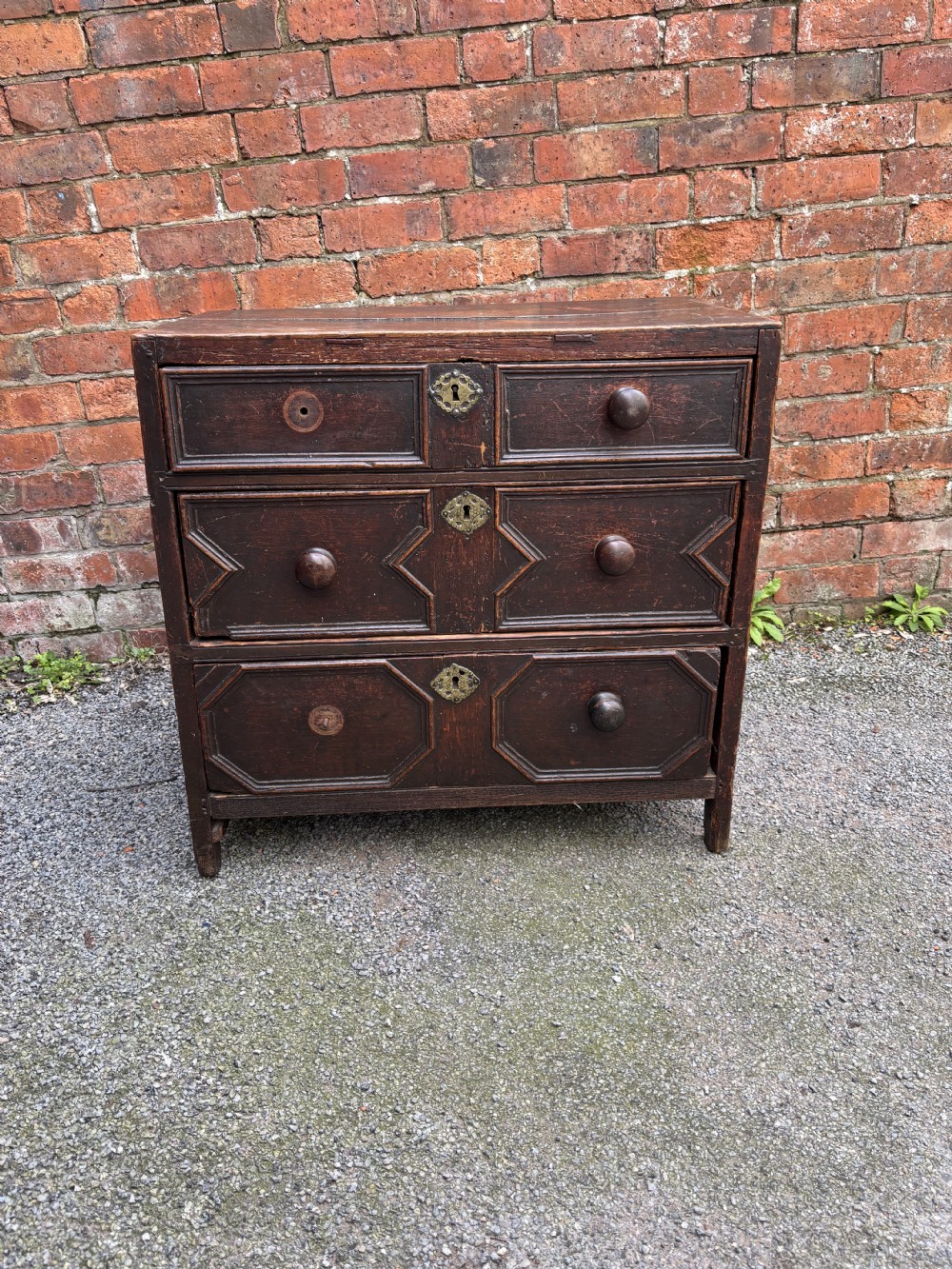 A dark oak 17th-century chest of drawers featuring geometric moulded drawer fronts, turned wooden handles, and brass escutcheons.