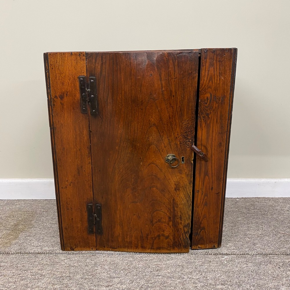 An early 18th-century rectangular warm brown oak hanging cupboard with a single timber door, hand-forged iron H-hinges, a brass ring handle, and a wooden latch.