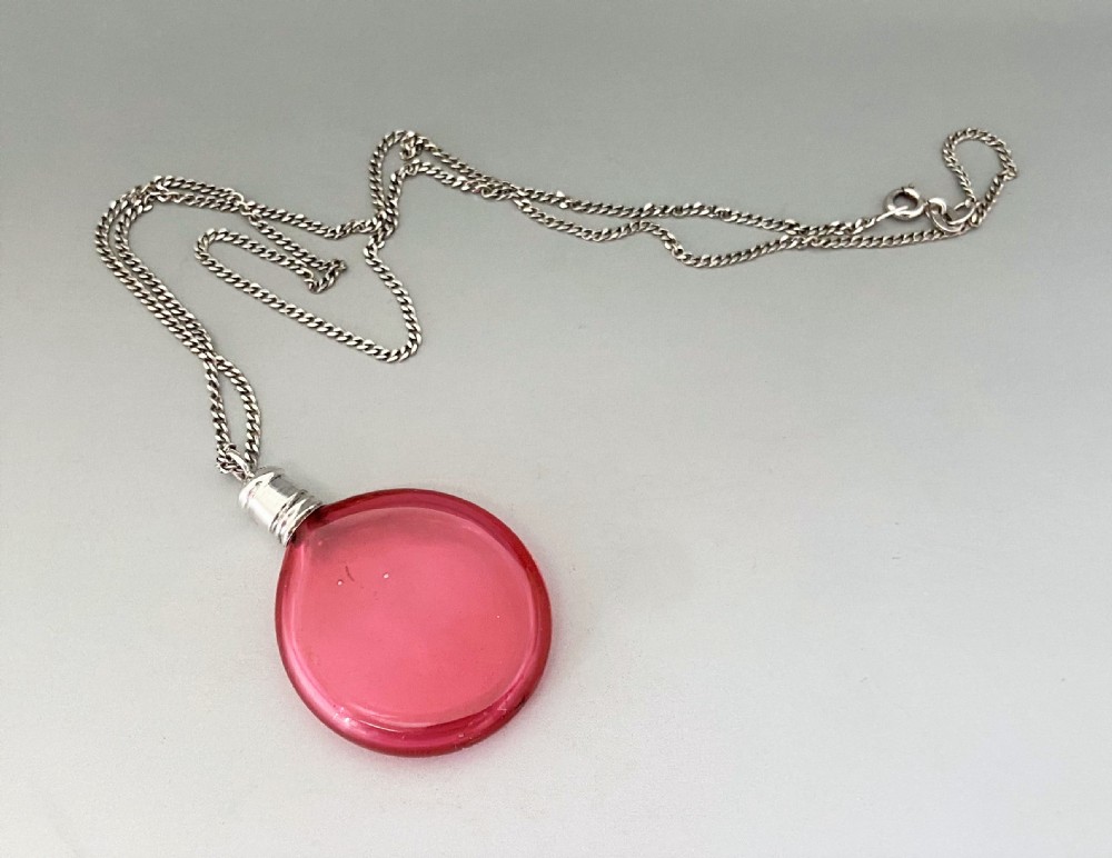 A Victorian circular cranberry glass penny scent bottle with a silver screw-top mount suspended on a fine silver chain. View II