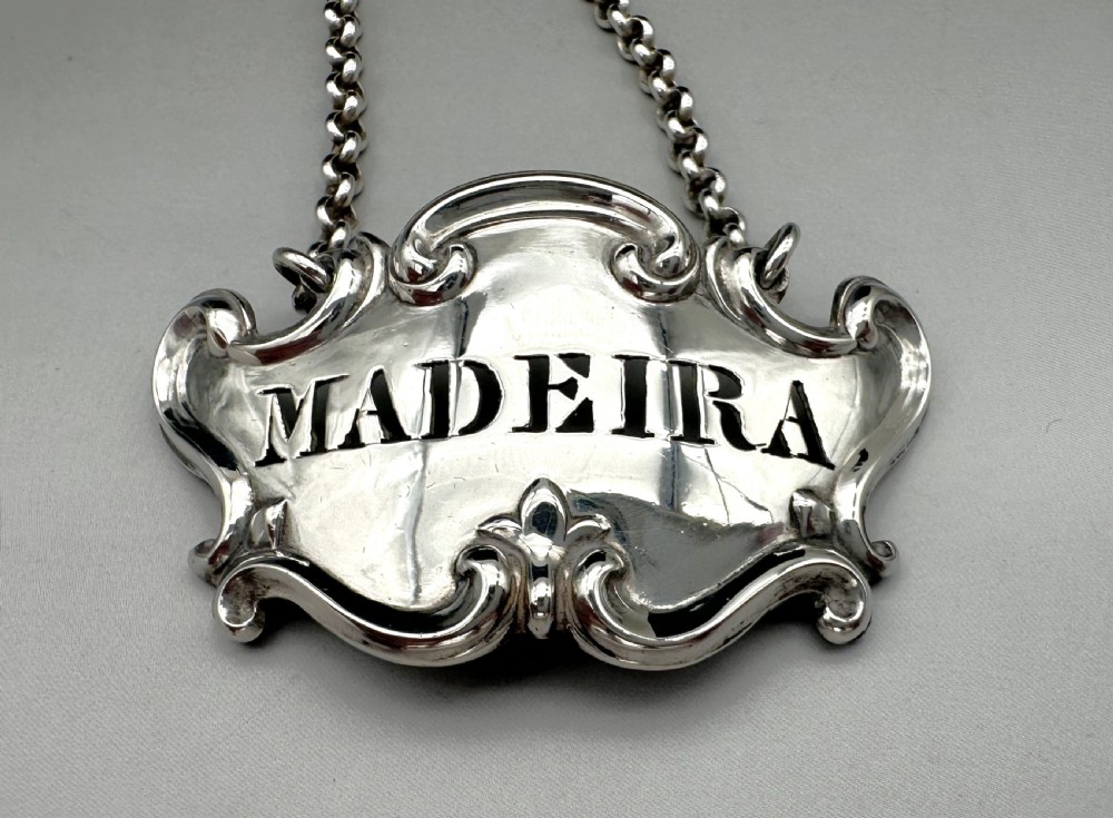 A Victorian sterling silver decanter label engraved with the word MADEIRA, featuring a decorative scrollwork border attached to a suspension chain. View II
