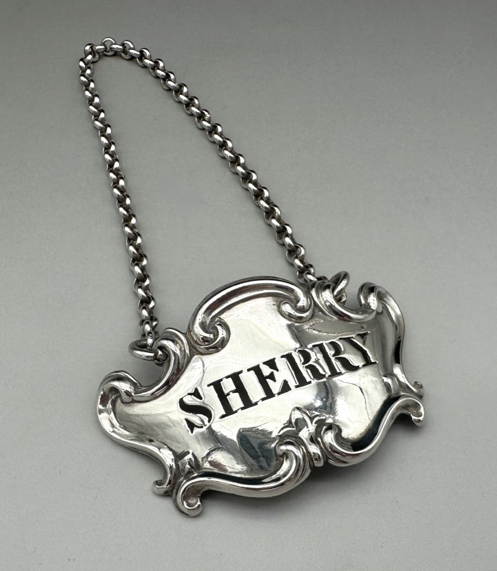 A Victorian sterling silver decanter label with intricate scrollwork borders and a suspension chain, featuring the word SHERRY cut into the centre.