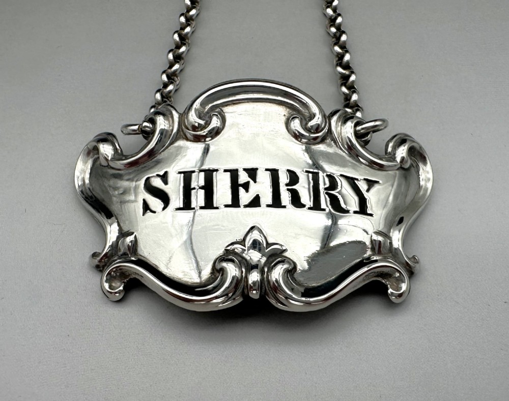 A Victorian sterling silver decanter label with intricate scrollwork borders and a suspension chain, featuring the word SHERRY cut into the centre. View II
