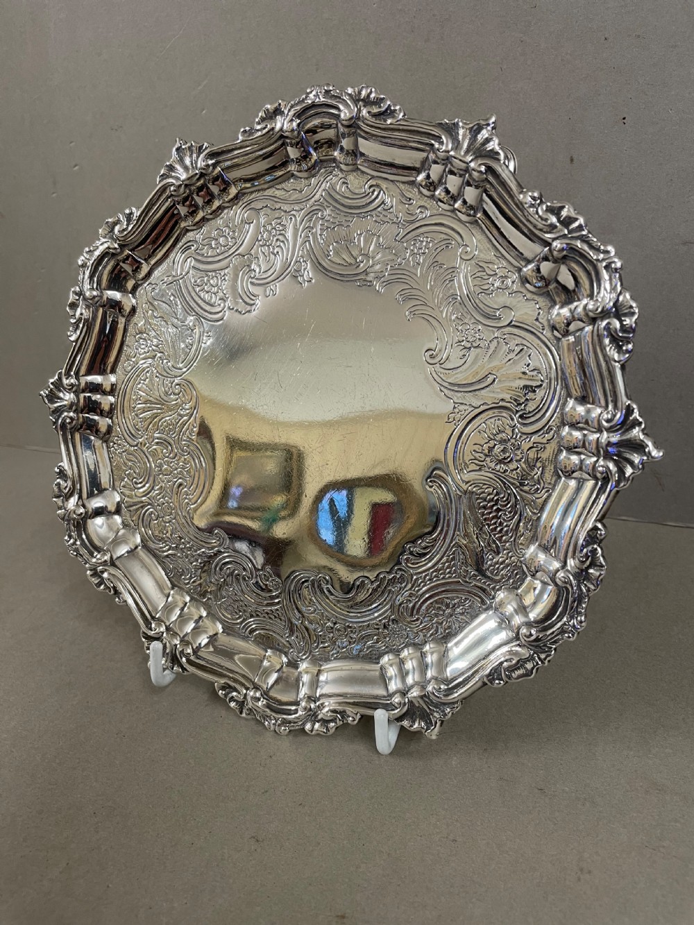 An ornate circular antique silver salver featuring heavily chased floral scrollwork and a raised scalloped rim.