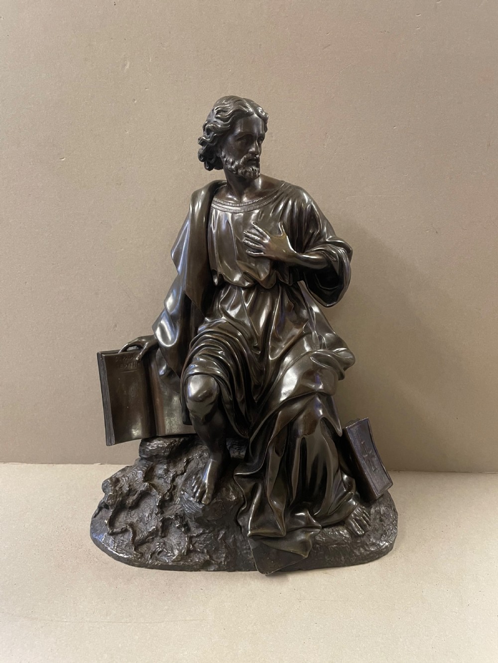 A dark bronze sculpture of a bearded male figure, identified as Saint Paul, kneeling amongst rocky terrain with books at his side.