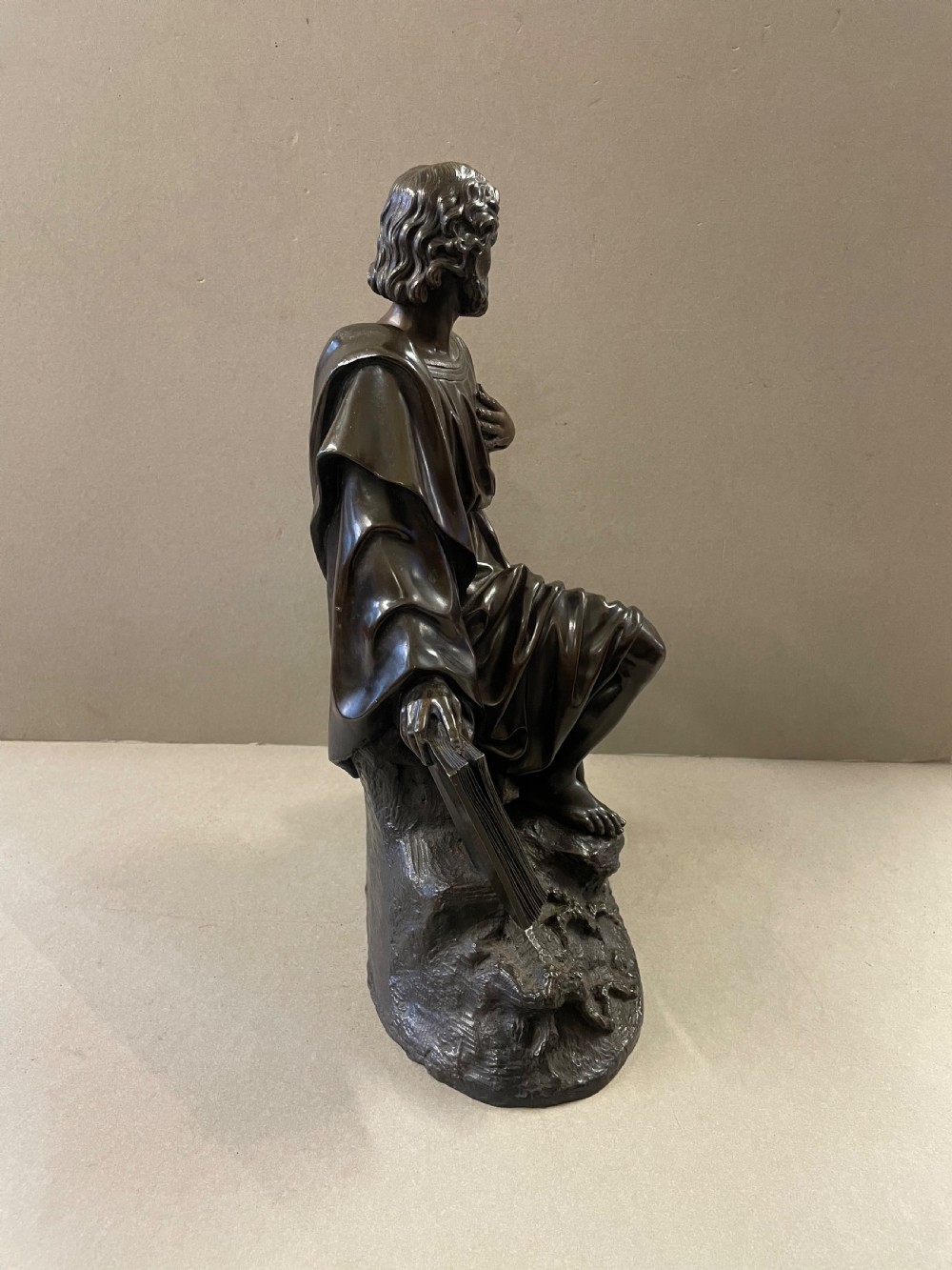 A dark bronze sculpture of a bearded male figure, identified as Saint Paul, kneeling amongst rocky terrain with books at his side. View IV