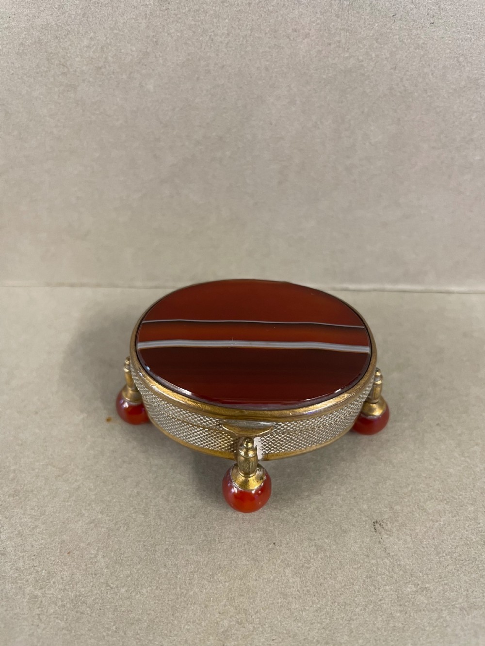 An antique small circular banded agate trinket box with reddish-brown and white stone panels, mounted in textured gilt metal with ball feet.