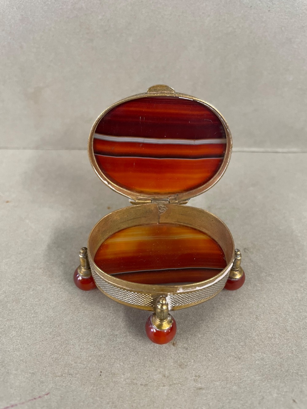 An antique small circular banded agate trinket box with reddish-brown and white stone panels, mounted in textured gilt metal with ball feet. View II