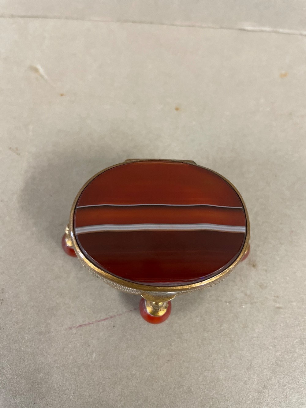 An antique small circular banded agate trinket box with reddish-brown and white stone panels, mounted in textured gilt metal with ball feet. View III