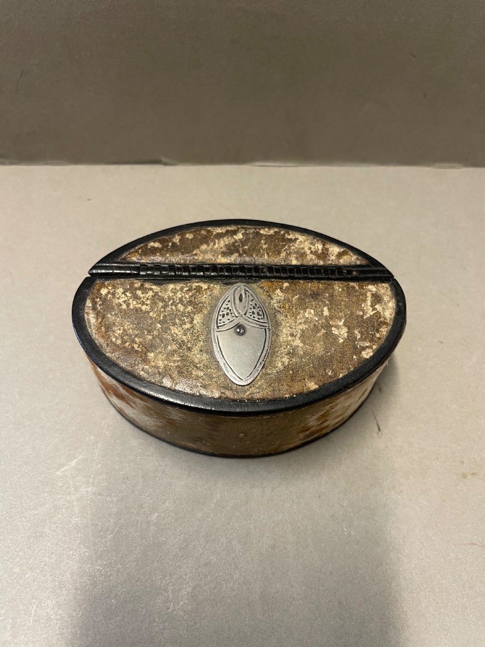 An oval antique snuff box featuring a mottled leather-covered body, a hinged lid, and an engraved metal escutcheon plate.