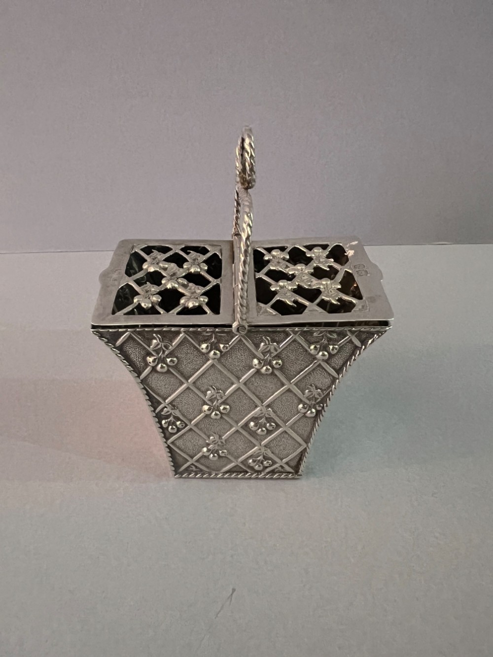 A small silver basket featuring double hinged pierced lids, a central rope-twist handle, and a diaper-patterned body with floral motifs.