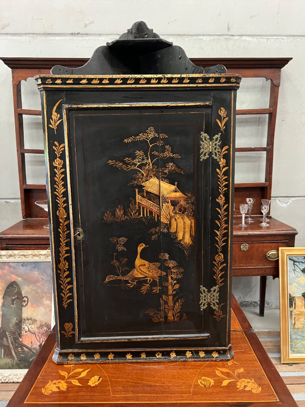 A dark black japanned hanging corner cabinet featuring gilt chinoiserie artwork with birds, architectural pagodas, and brass hinges.