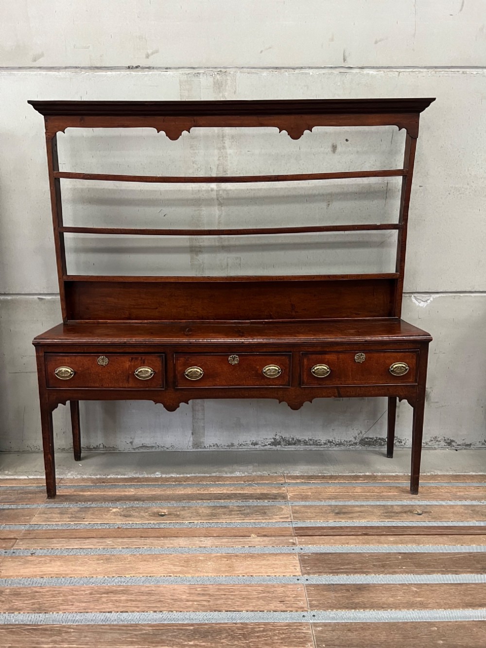 An 18th century oak and elm Welsh dresser featuring an open three-tiered plate rack top above three drawers with brass handles and a shaped apron.