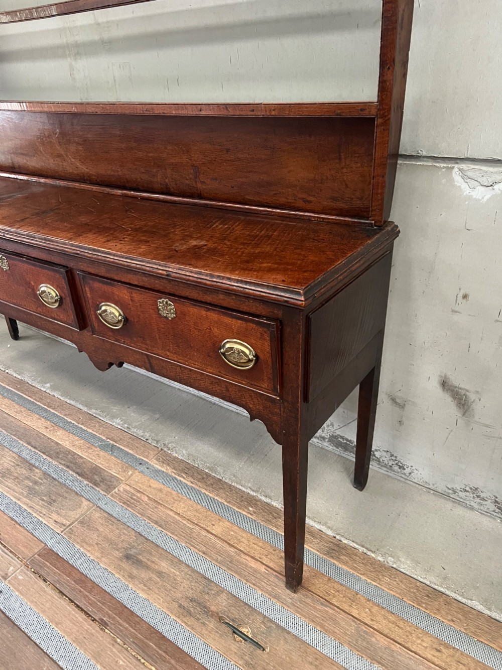 An 18th century oak and elm Welsh dresser featuring an open three-tiered plate rack top above three drawers with brass handles and a shaped apron. View V